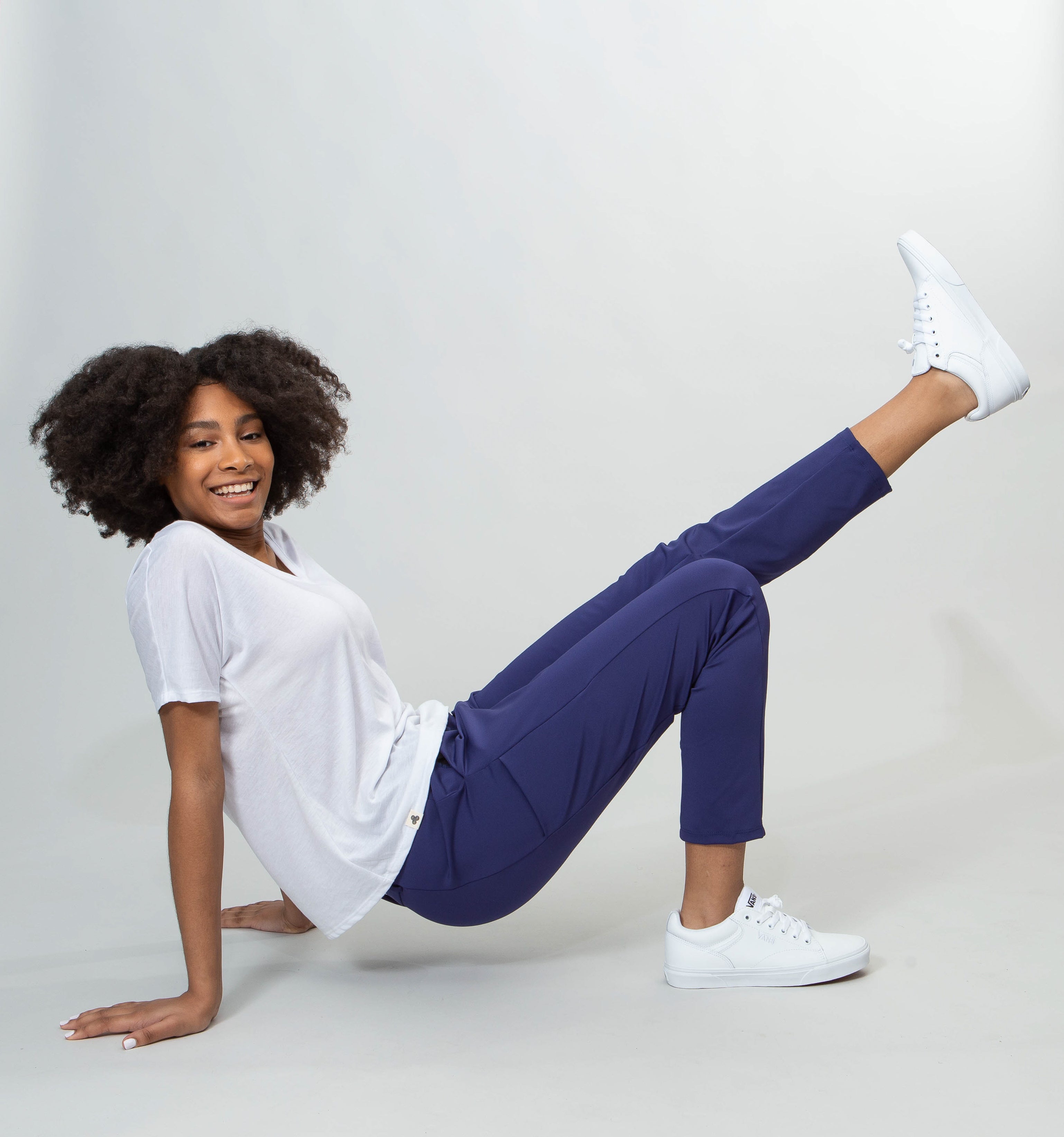 African American model smiling at us, postured on the ground arms behind and palms down, one leg up in the air.  She has a white shirt and blue pants.